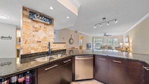 A modern kitchen with a stone backsplash, a built-in dishwasher, and a large island with a sink. - Gulf & Bay Club Bayside | Walk to Beach, Enjoy Pools, Tennis & More (Siesta Key)