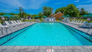 A rectangular swimming pool with a tiled edge, surrounded by lounge chairs and umbrellas, with a fenced area and a building in the background. - Brewster Green Resort  (Brewster)