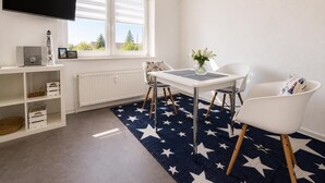 A room with a white table, two chairs, a TV, a radiator, and a window with blinds. - House Najade, small feel-good oasis Whg. 7, Pastoratsweg 17, 27476 Cuxhaven Döse (Cuxhaven)