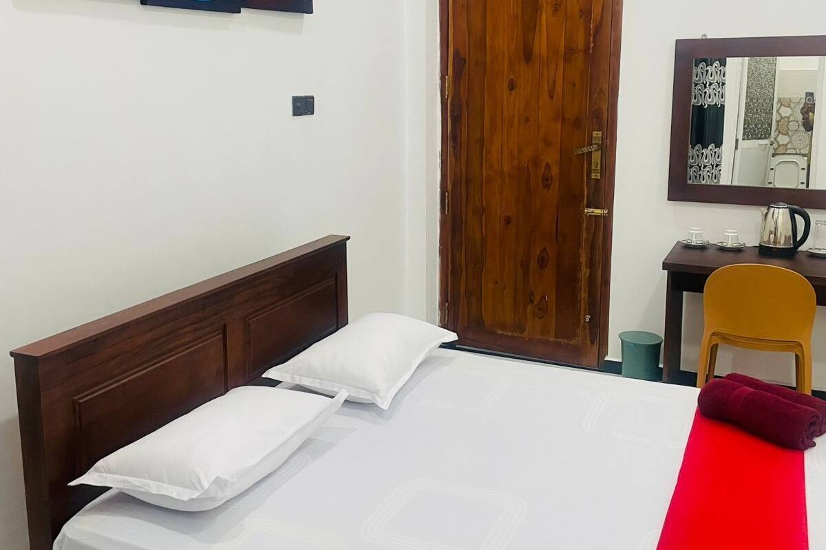 A bedroom with a wooden bed, a red bedspread, a wooden door, a desk with a chair, and a wall-mounted mirror.