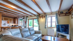 Ein großzügiger Wohnraum mit einer großen, lederbezogenen L-Form-Couch, einem Holztisch und einem Fernseher auf einem Schrank. Der Raum zeichnet sich durch freiliegende Holzbalken an der Decke und große Fenster mit Vorhängen aus.