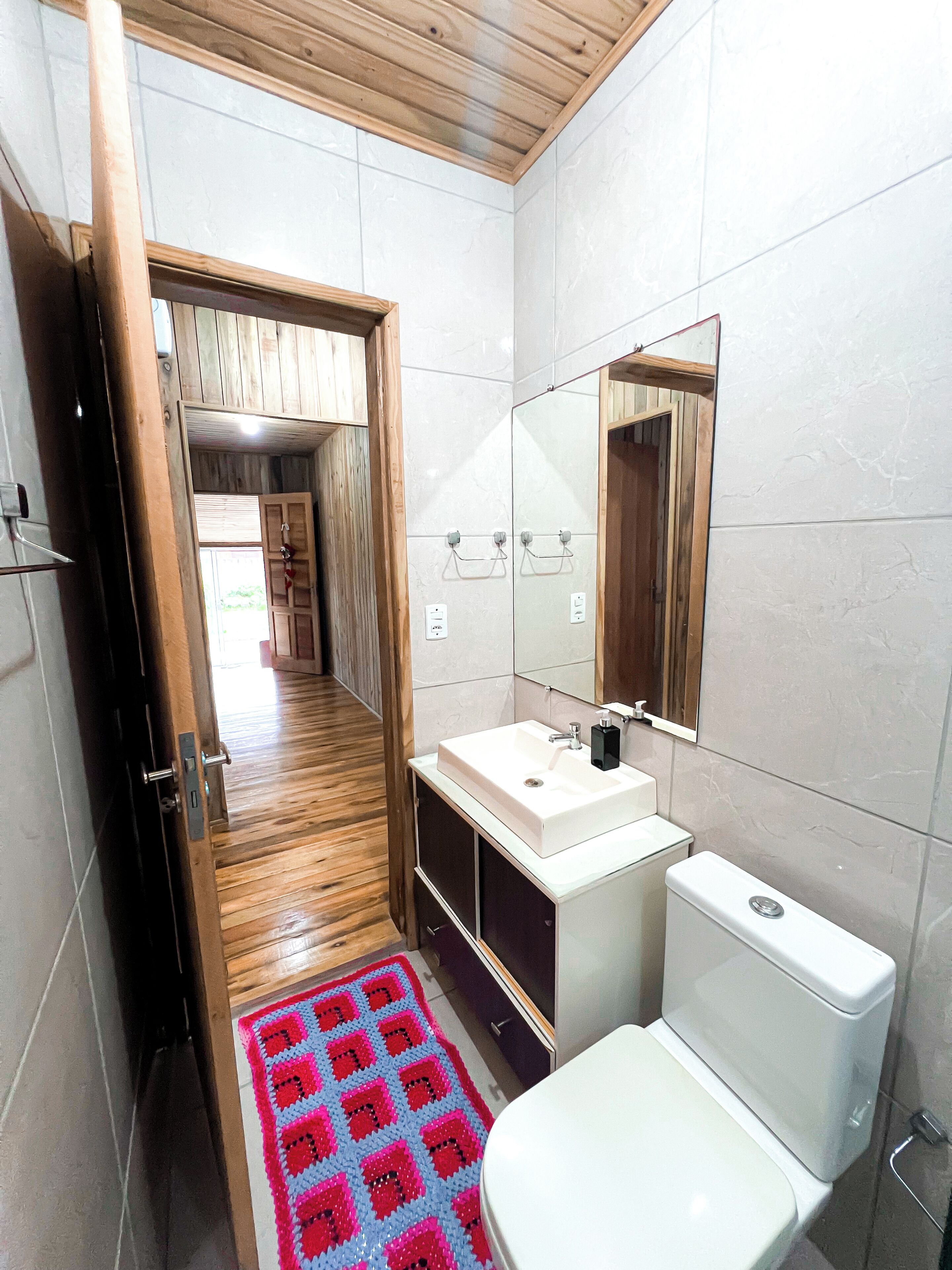 A narrow bathroom with a wooden floor, white tiles, a white sink, a toilet, and a mirror.