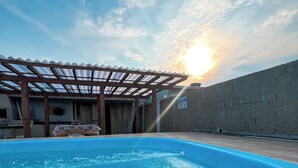 A swimming pool with a pergola overhead, a brick wall, and a sunset in the background.