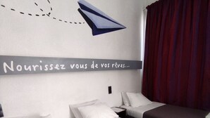 Une chambre d’hôtel avec deux lits, un mur sur lequel est écrit un message en français, un rideau rouge et une serviette pliée sur le sol.