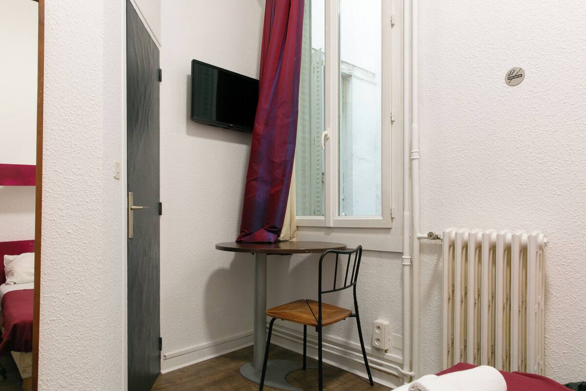 Ein kleines Zimmer mit einem Bett, einem Tisch mit Stuhl, einem an der Wand befestigten Fernseher und einem Fenster mit Vorhängen.