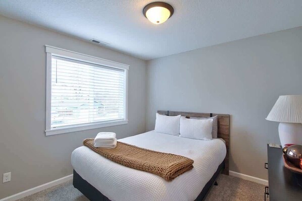 A neatly made bed with white linens and a brown throw blanket, a bedside lamp, and a window with blinds.