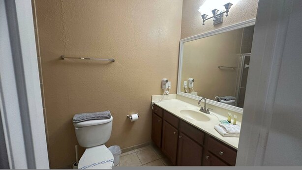 A bathroom with a beige wall, a white toilet, a sink with a mirror, and a towel rack. - Cozy Condo Just a few Minutes From Disney (Kissimmee)