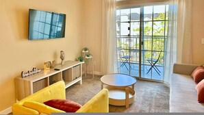 Un salon doté d’un téléviseur à écran plat fixé au mur, d’une table basse ronde, de chaises jaunes et rouges, d’un canapé et d’une porte-fenêtre coulissante donnant sur un balcon extérieur avec une table et des chaises.