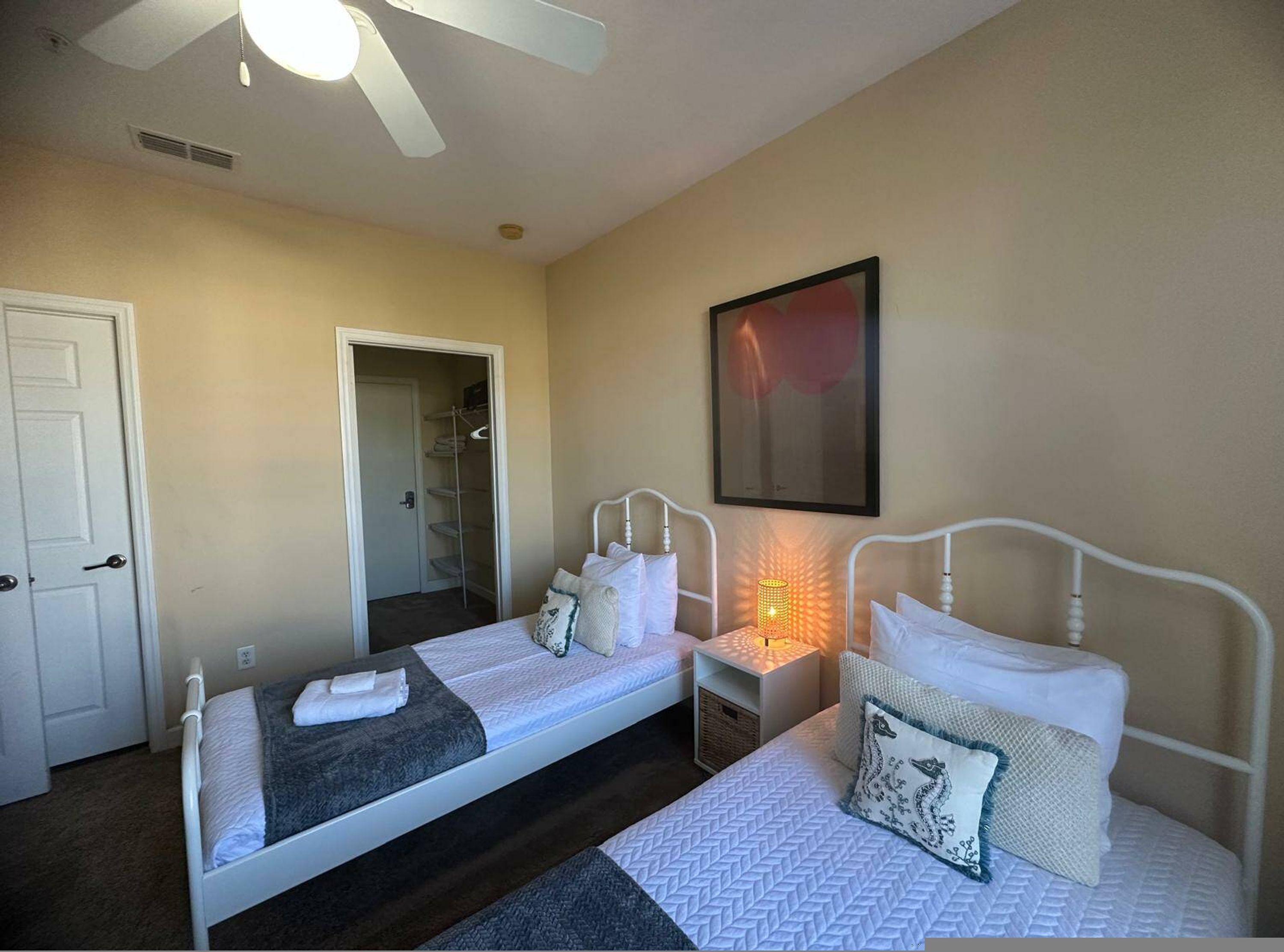 A room with two single beds, a ceiling fan, a door, a framed picture, and a small table with a lamp.