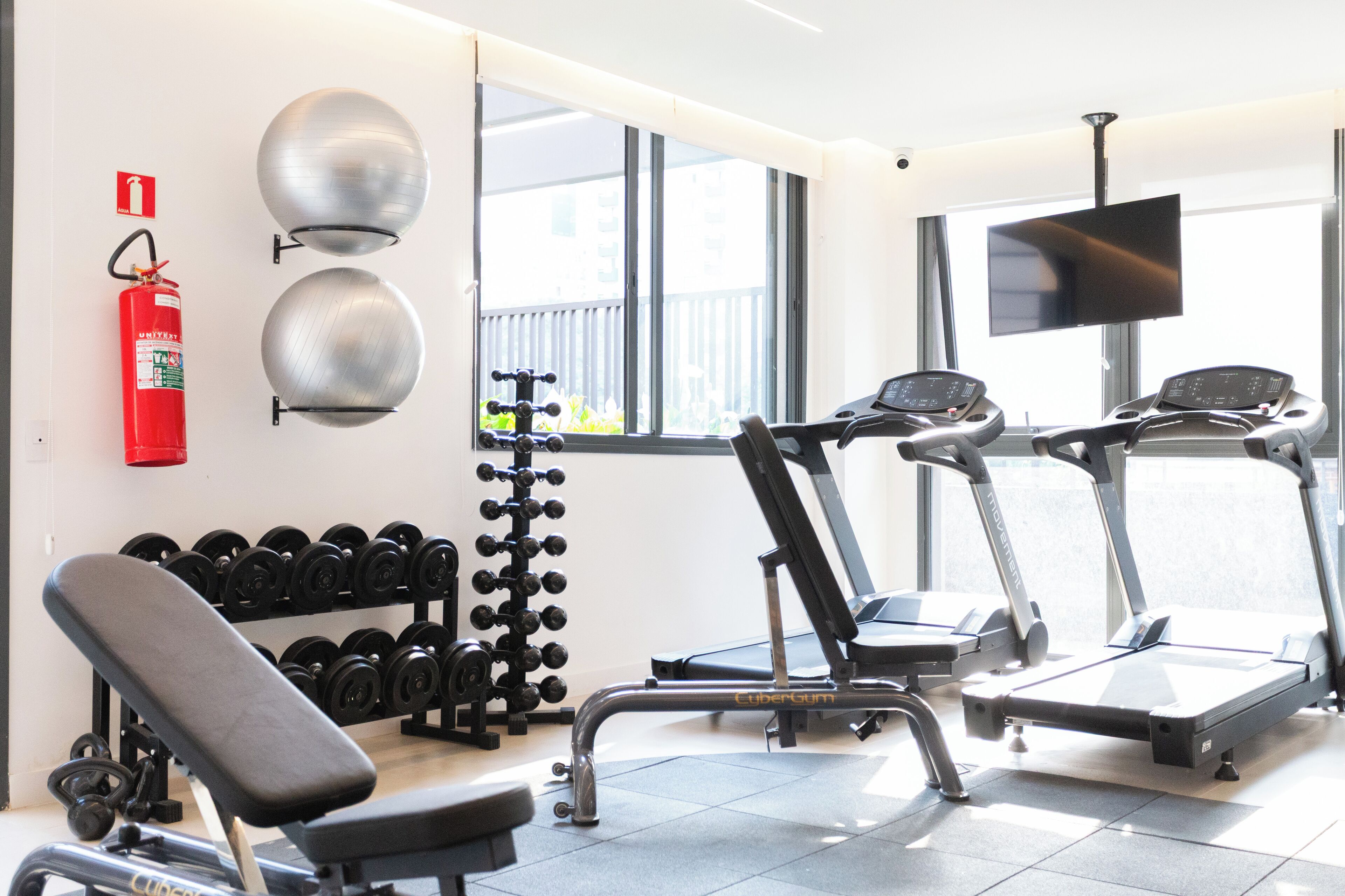 A gym with exercise equipment, a fire extinguisher, and a TV on the wall.