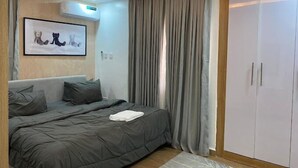A modern bedroom with a bed, grey bedding, a framed artwork, a wardrobe, and a window with curtains. - Amazon Luxury Apartment (Enugu)