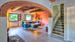 A spacious living area with a modern sofa set, a blue armchair, and a large window providing natural light. A staircase with a stone wall and a metal railing is visible to the right. - Tuscan villa for 8 people with pool near Lucca. (Borgo a Mozzano)
