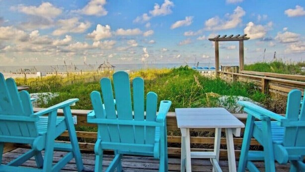 Een houten terras met turquoise Adirondack-stoelen en een kleine tafel, met uitzicht op een strand met een houten pier en een prieeltje.