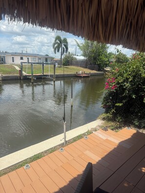 A wooden deck with a fishing rod, a concrete path, and a water body with a fence and greenery.
