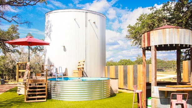 Un jardin avec un grand réservoir d’eau blanche, une petite piscine, des structures de jeux en bois et un belvédère rustique.