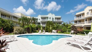 A resort pool with a 4.6' depth, surrounded by lounge chairs and umbrellas, with a building featuring balconies in the background. - Charming 2-bedroom condo in fabulous Hastings area (Hastings)