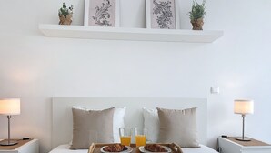 A neatly made bed with a tray of croissants and orange juice, flanked by two bedside lamps, against a white wall with decorative elements above. - Acanto Loft by Atlantic Holiday (Funchal)