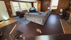 A cozy living room with a sectional sofa, a flat-screen TV, wooden floors, and large windows.