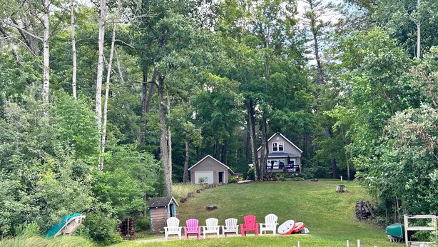 A lakeside cabin with a dock, surrounded by trees and a grassy area with Adirondack chairs.