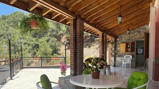 An outdoor patio area with a round table and green cushioned chairs, a brick wall, and a stone staircase leading to a kitchen area. - VILLA AURA (Πλάτρες)
