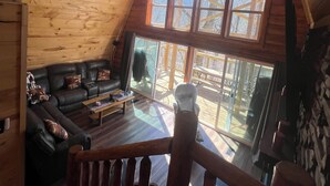 A cozy cabin interior with a wooden ceiling, a ceiling fan, a large window, a leather sofa, and a wooden deck visible through the window. - Cabin in the TreeTops! 4 Level a-frame cabin, with 5 levels of decks (Lancaster)