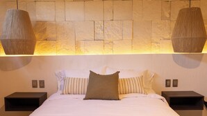 A neatly made bed with a white duvet and brown pillows, two bedside tables, and a stone wall with a warm light fixture above. - Traspatio hotel Boutique  (Bernal)