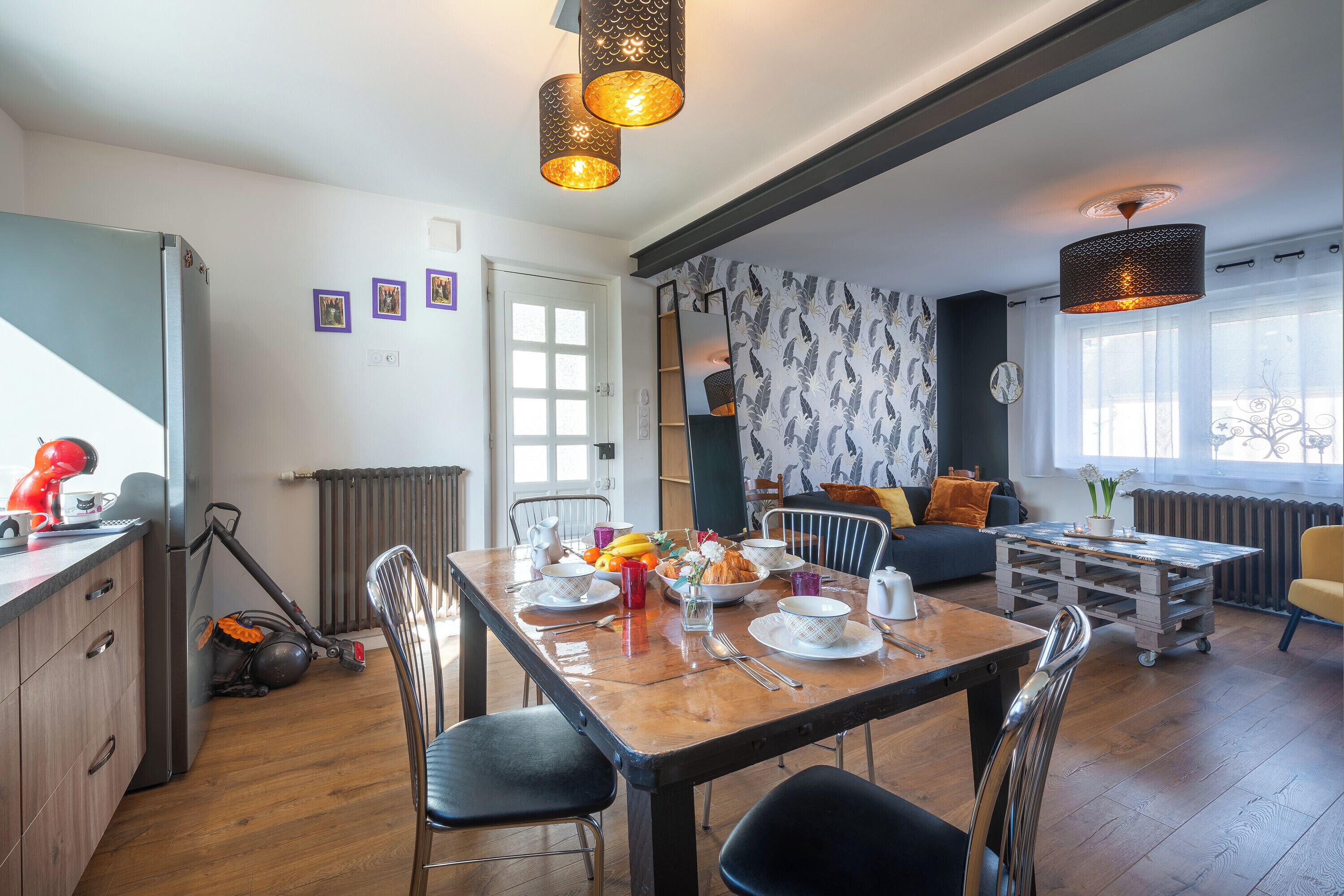 A dining area with a wooden table, chairs, and a variety of dishes. There is a radiator, a sofa, and a window with curtains.