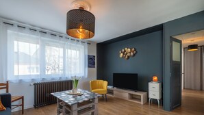 A modern living room with a wooden floor, a table with a patterned tablecloth, a yellow armchair, a TV, and a decorative wall piece. - Le Jovia - Cour extérieure - Wifi (Laval)