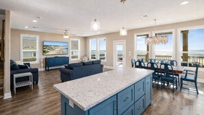 A spacious living area with a large island, a dining area with blue chairs, and a view of the ocean through large windows. - Beachfront | Hot Tub | Putt Putt | Fire Pit (Gilchrist)