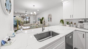 Una cocina moderna con armarios blancos, una isla grande con pileta y un comedor visible a través de un ventanal.