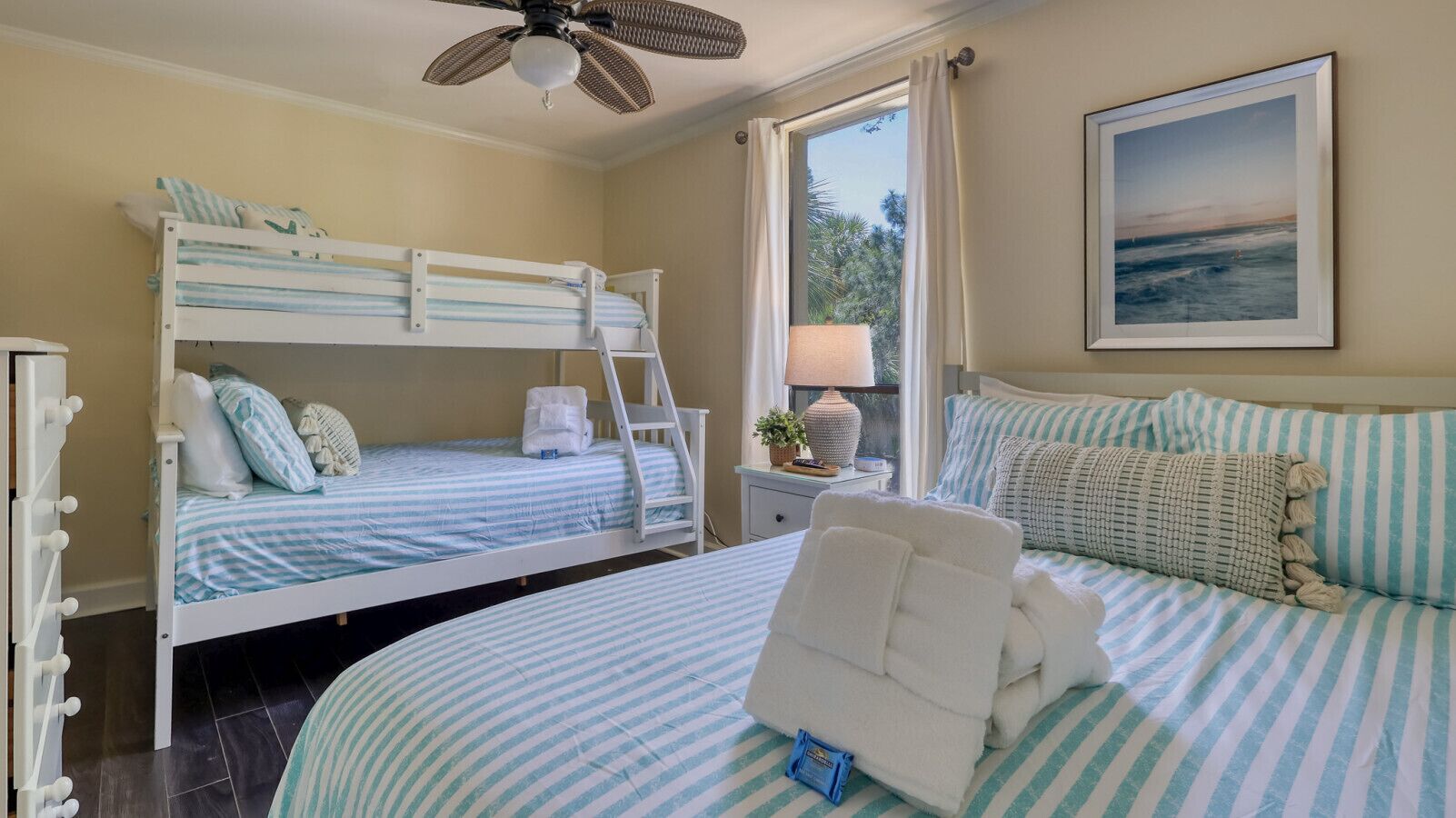 A bedroom with two bunk beds, a large bed with striped bedding, a ceiling fan, a window with curtains, and a framed picture on the wall.