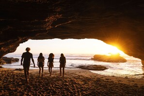 A group of people walking on a beach with a cave entrance and the sun setting in the background.