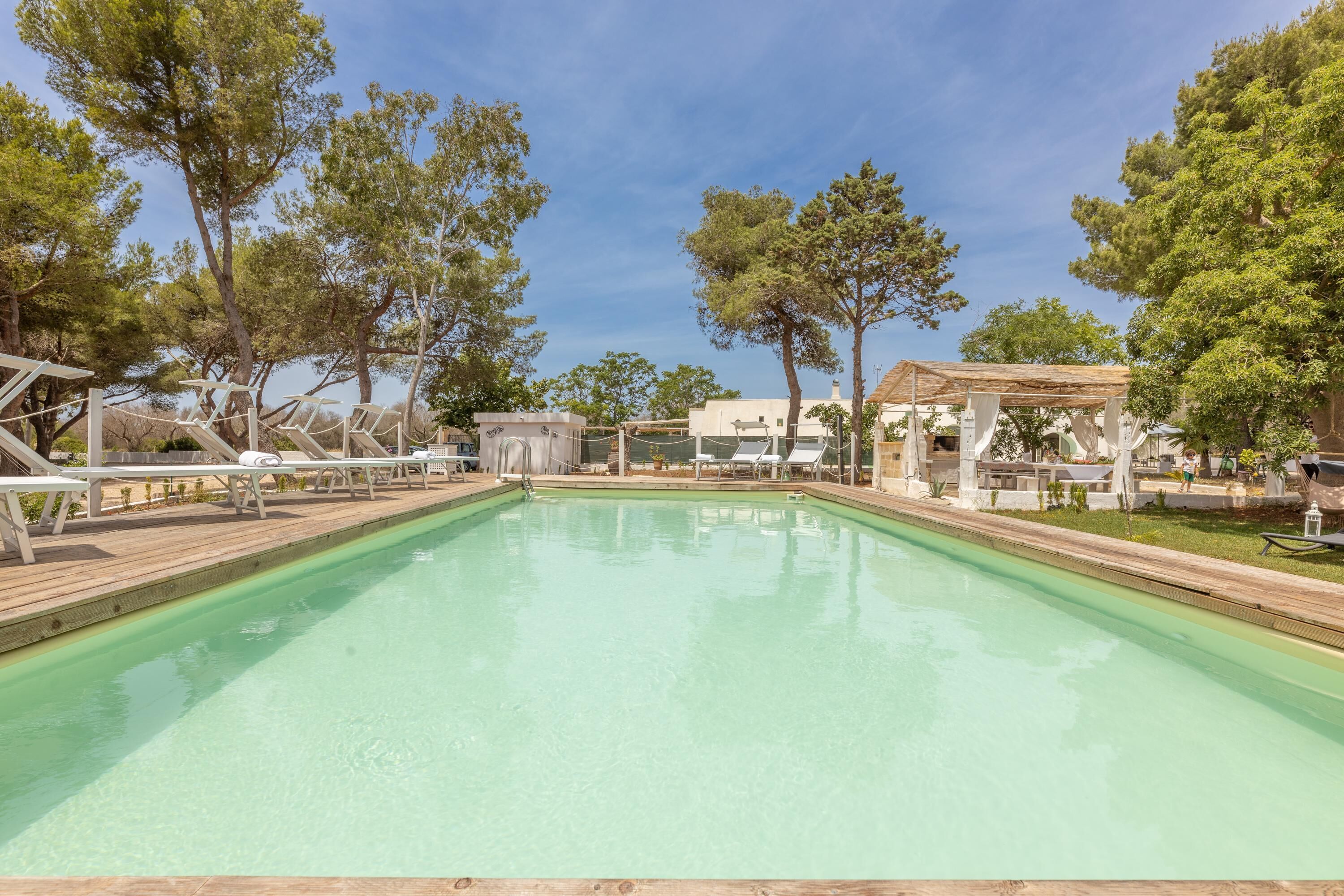 A rectangular swimming pool with a wooden deck, surrounded by trees and shaded areas with lounge chairs.