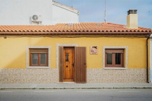 A yellow building with a tiled roof, a wooden door, and two windows with shutters.