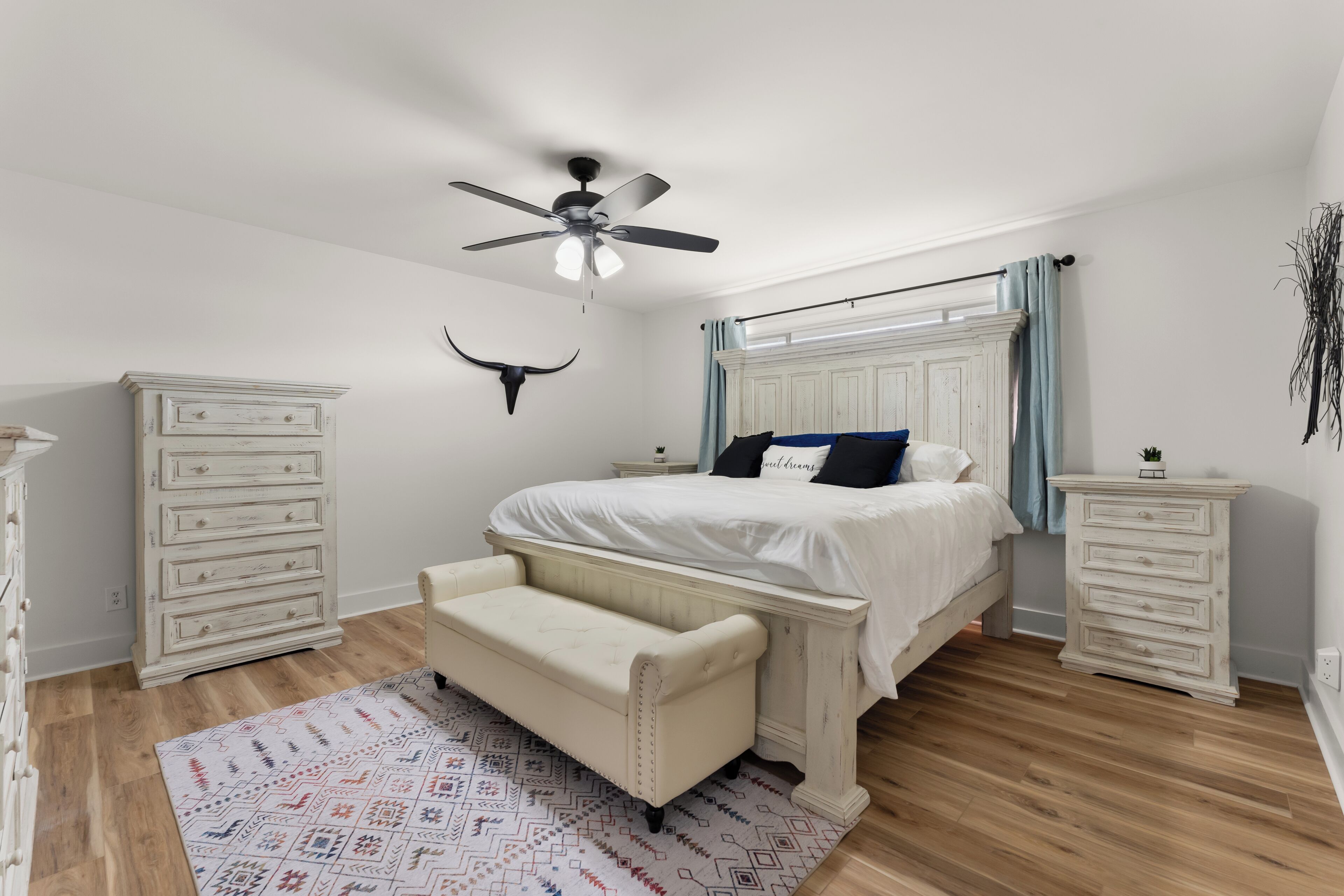 Ein Schlafzimmer mit einem Bett, einer Bank, einem Schminktisch, einem Nachttisch, einem Deckenventilator und einer Wanddekoration.