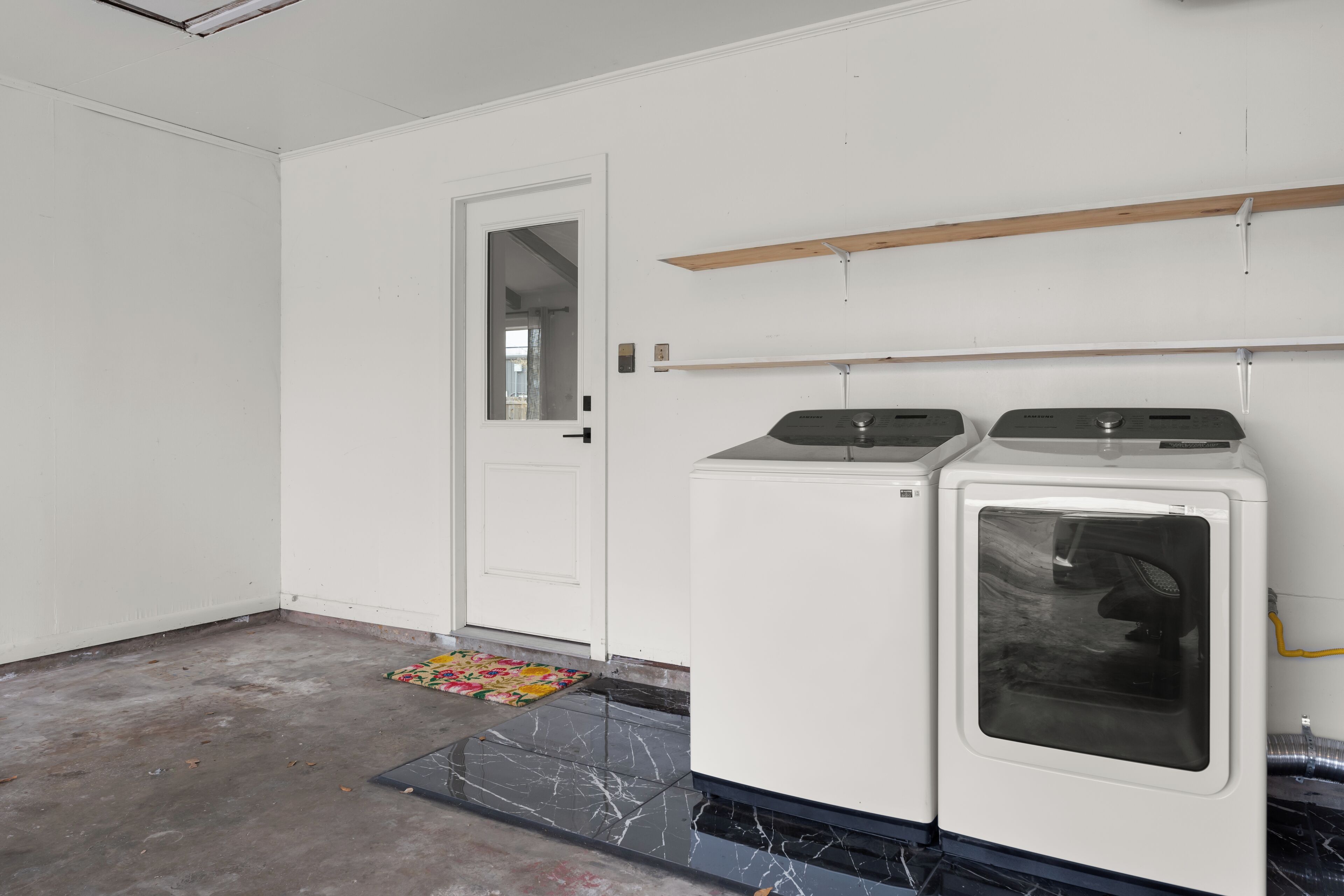 A laundry room with two washing machines, a door, and a wooden shelf above the machines.