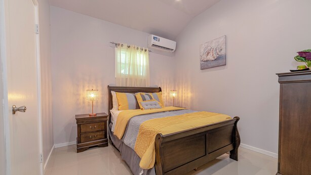A bedroom with a wooden bed, bedside tables, a nightstand, a wall-mounted air conditioner, a window with curtains, and a painting on the wall. - Beautiful 1-bedroom apartment in amazing Buckleys with AC, WiFi (Buckleys)