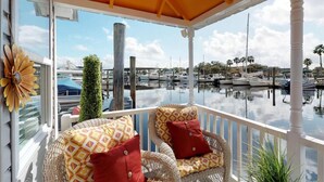 A covered balcony with wicker furniture and red cushions, overlooking a marina with boats and palm trees.