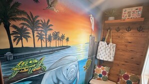 A room with a mural of a sunset, a wooden shelf with decorative items, a bench with colorful cushions, and a wall-mounted plant.