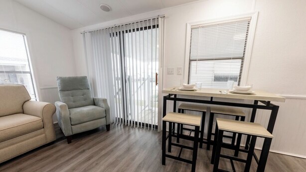 A modern living room with a beige sofa, a grey armchair, a wooden dining table with two chairs, and a window with blinds. - Fully remodeled cozy mobile home in charming Zephyrhills with AC, WiFi (Zephyrhills)
