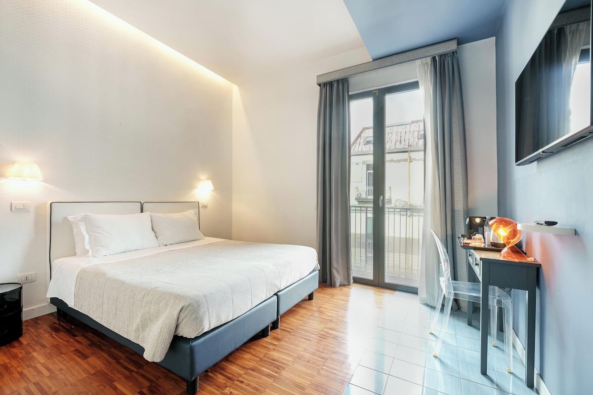 A modern hotel room with a bed, a desk with a lamp, a chair, and a balcony with a view.
