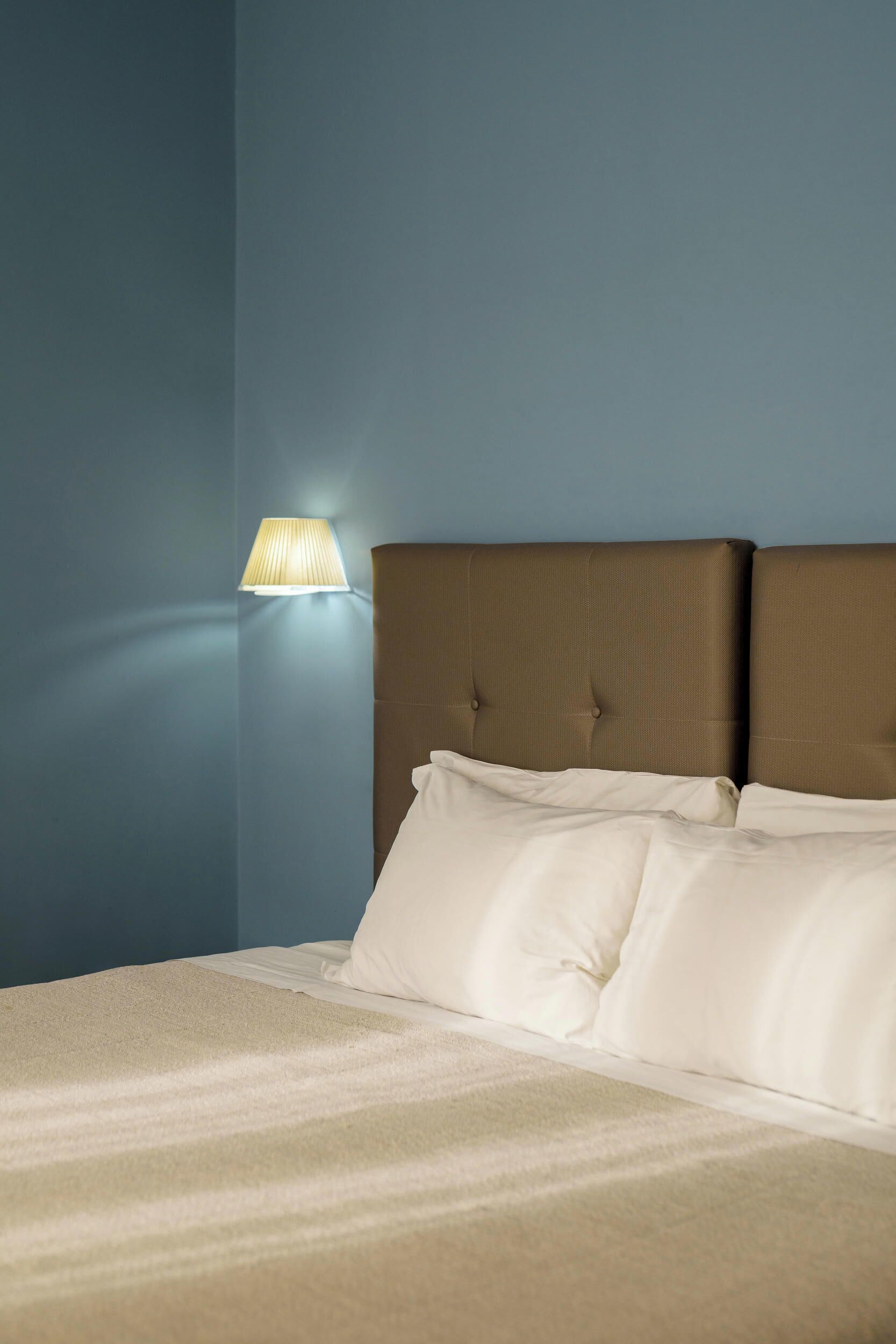 A neatly made bed with white linens in a room with blue walls and a wall-mounted lamp.