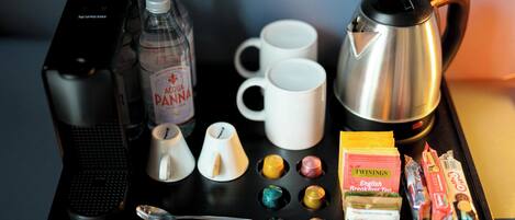 A coffee station with a coffee maker, cups, a kettle, a bottle of water, and various breakfast items.