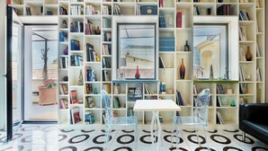 Une pièce comprenant une grande bibliothèque remplie de livres, une chaise en plastique transparent, une table blanche et un canapé noir.