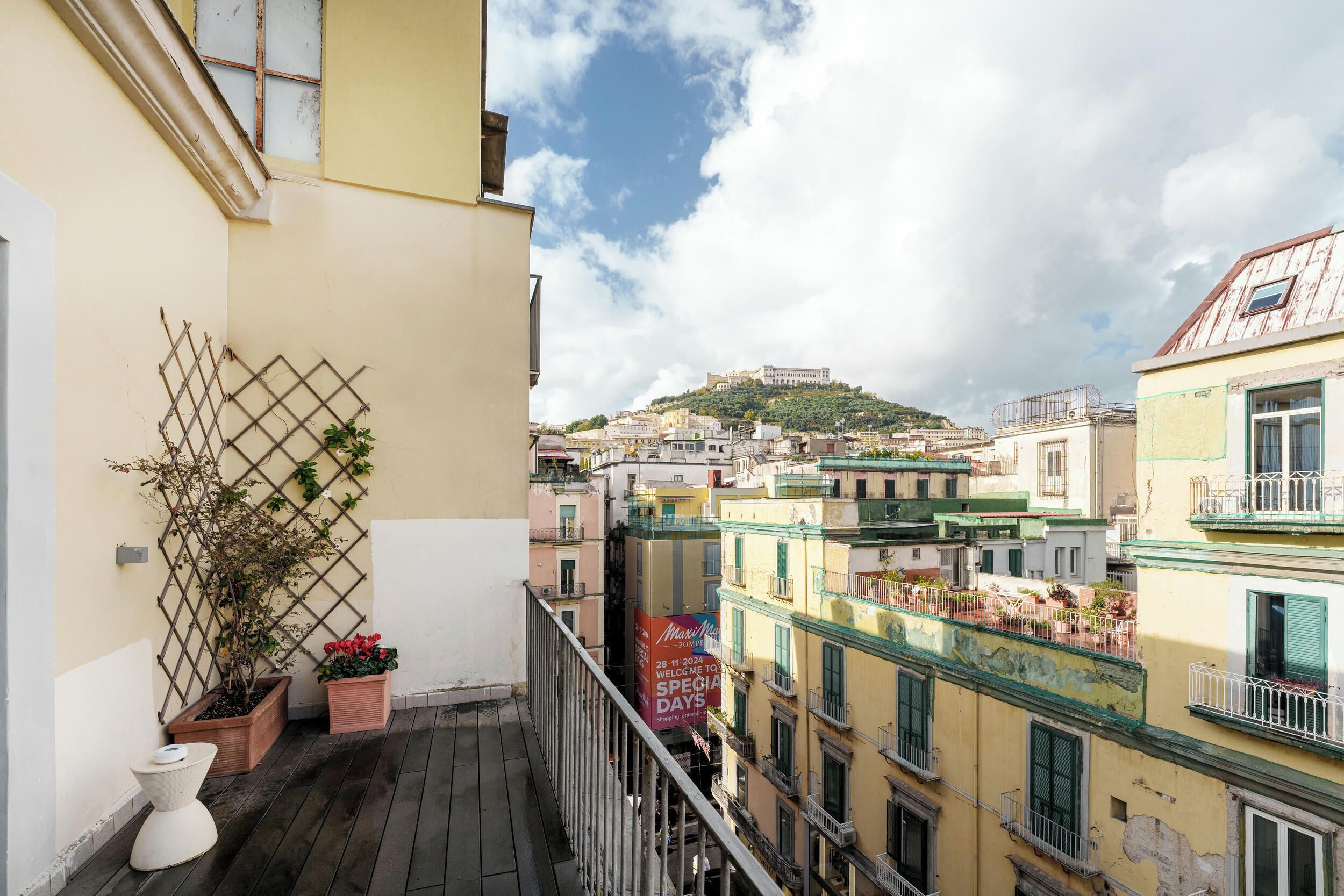 Un balcón con celosía, plantas en macetas y vista a una colina con edificios.