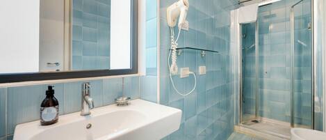 Un baño con lavamanos blanco, secador de pelo de mano instalado en la pared, cabina de ducha de vidrio y un inodoro.