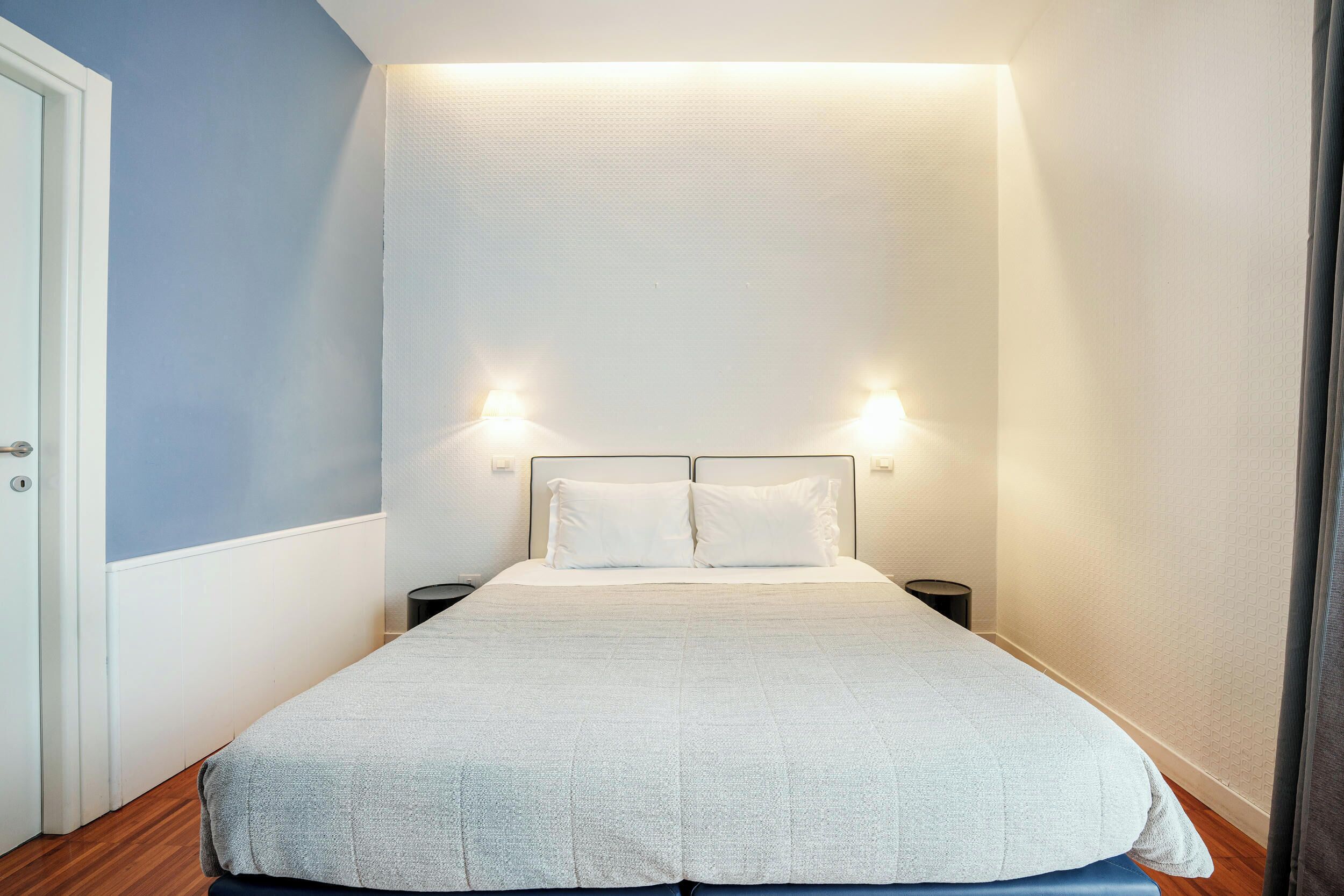 A neatly made bed with a white duvet and pillows, two bedside tables with lamps, and a door to the left.