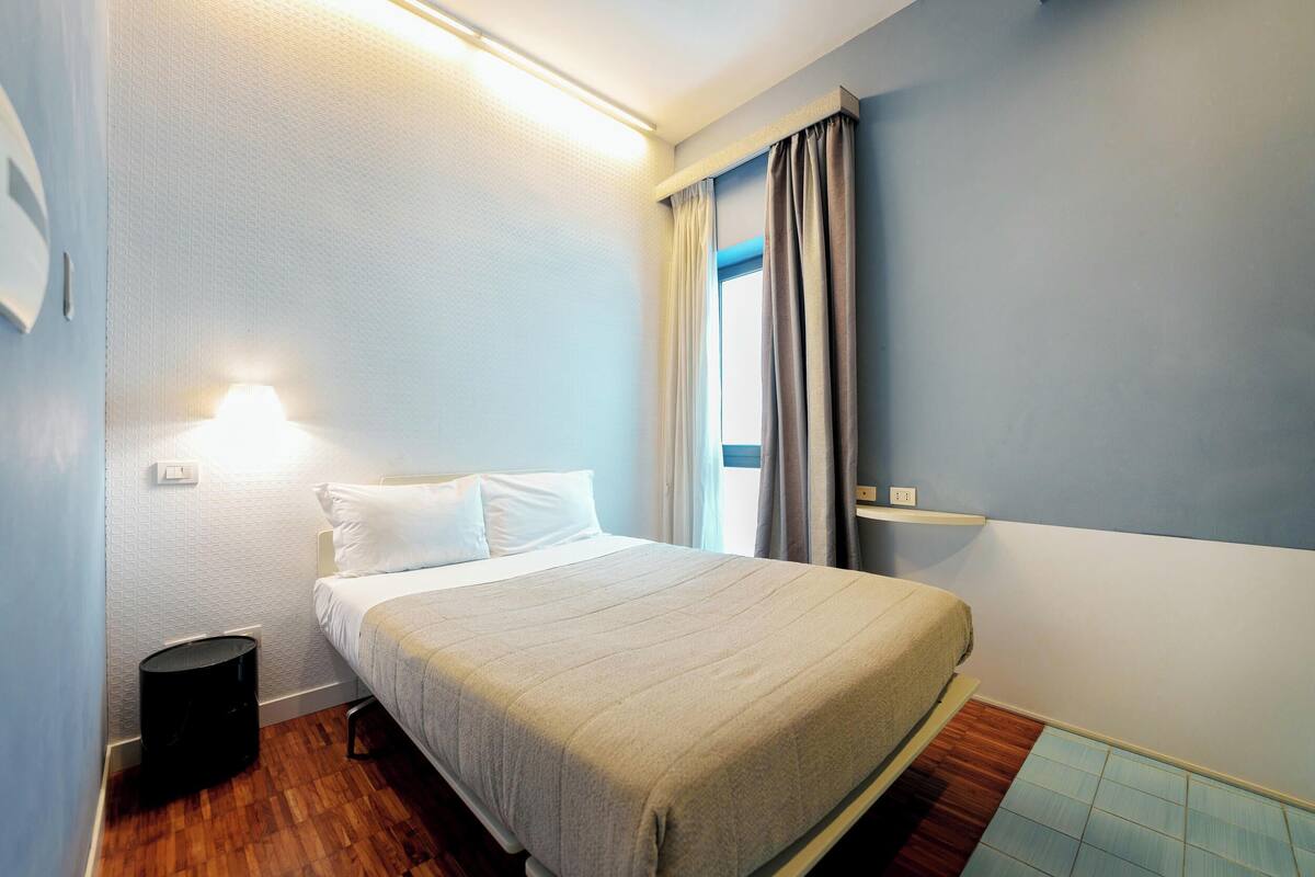 A neatly made bed in a room with a wooden floor, a small black trash bin, and a window with curtains.