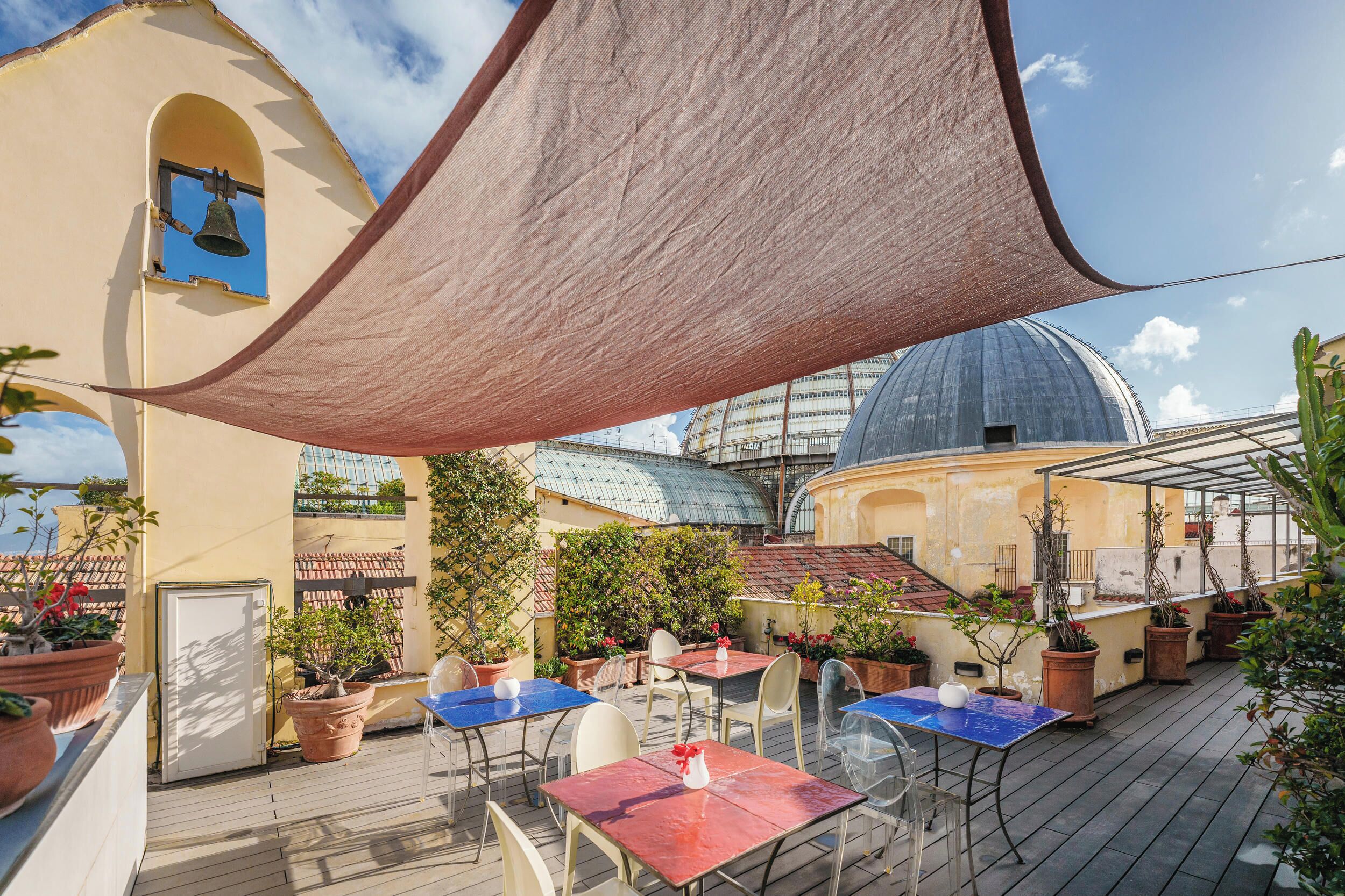 An outdoor patio with tables and chairs, surrounded by potted plants and a bell tower.