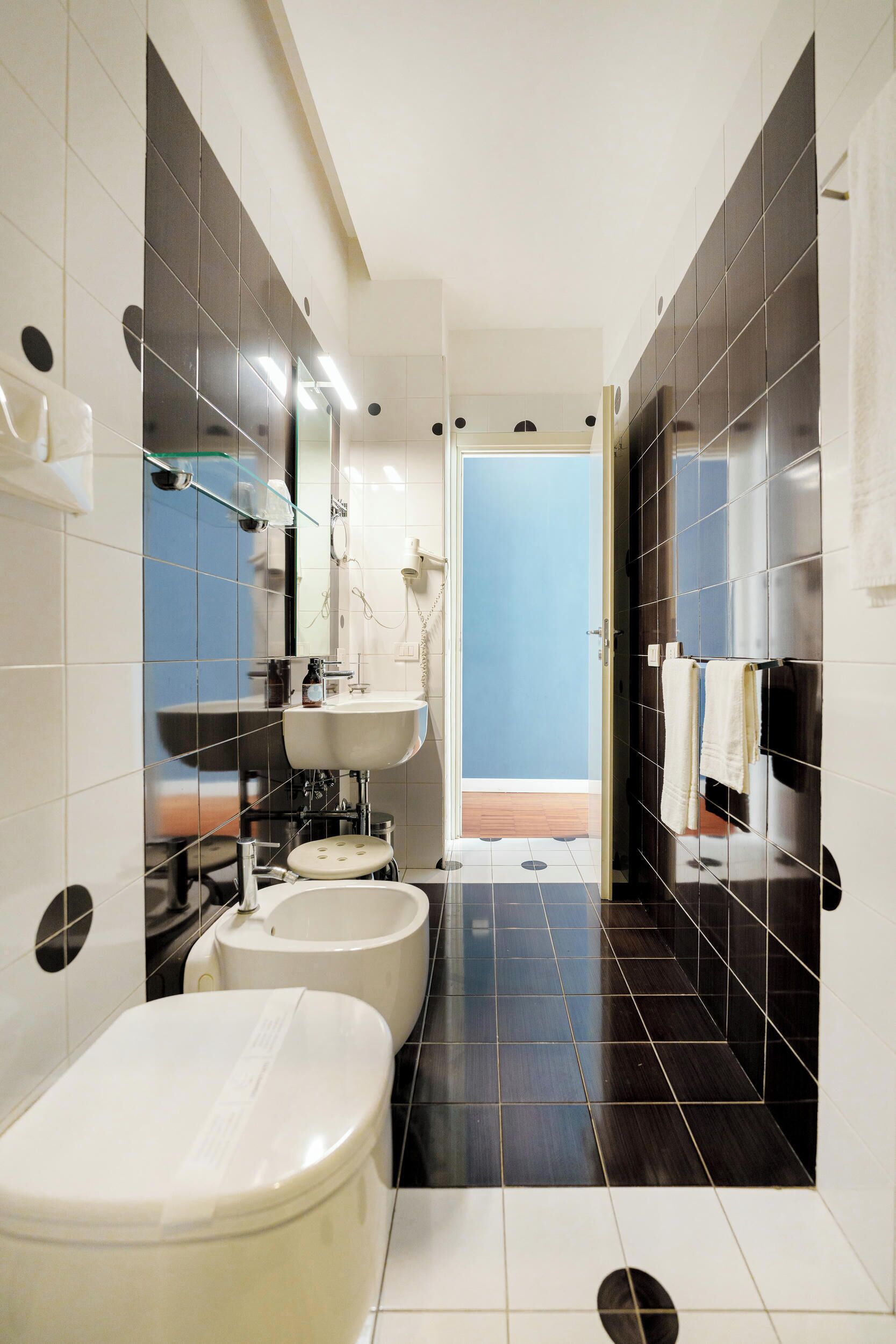A modern bathroom with a toilet, bidet, and sink, featuring dark and light tiles, a mirror, and a towel rack.